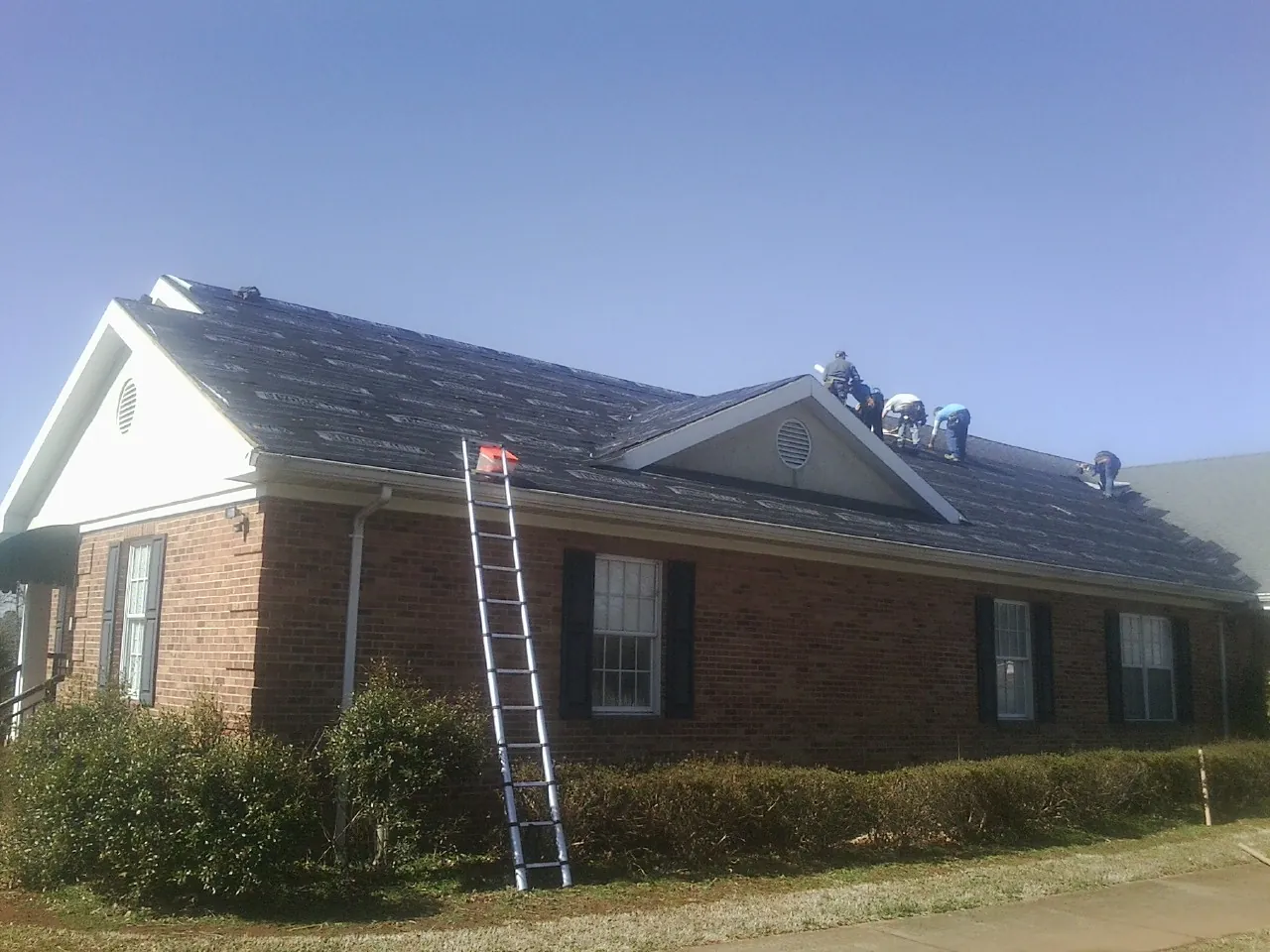 Crew tearing shingles off a brick ranch roof with a ladder set — Emergency Roof Repair work by our crew in Jessie
