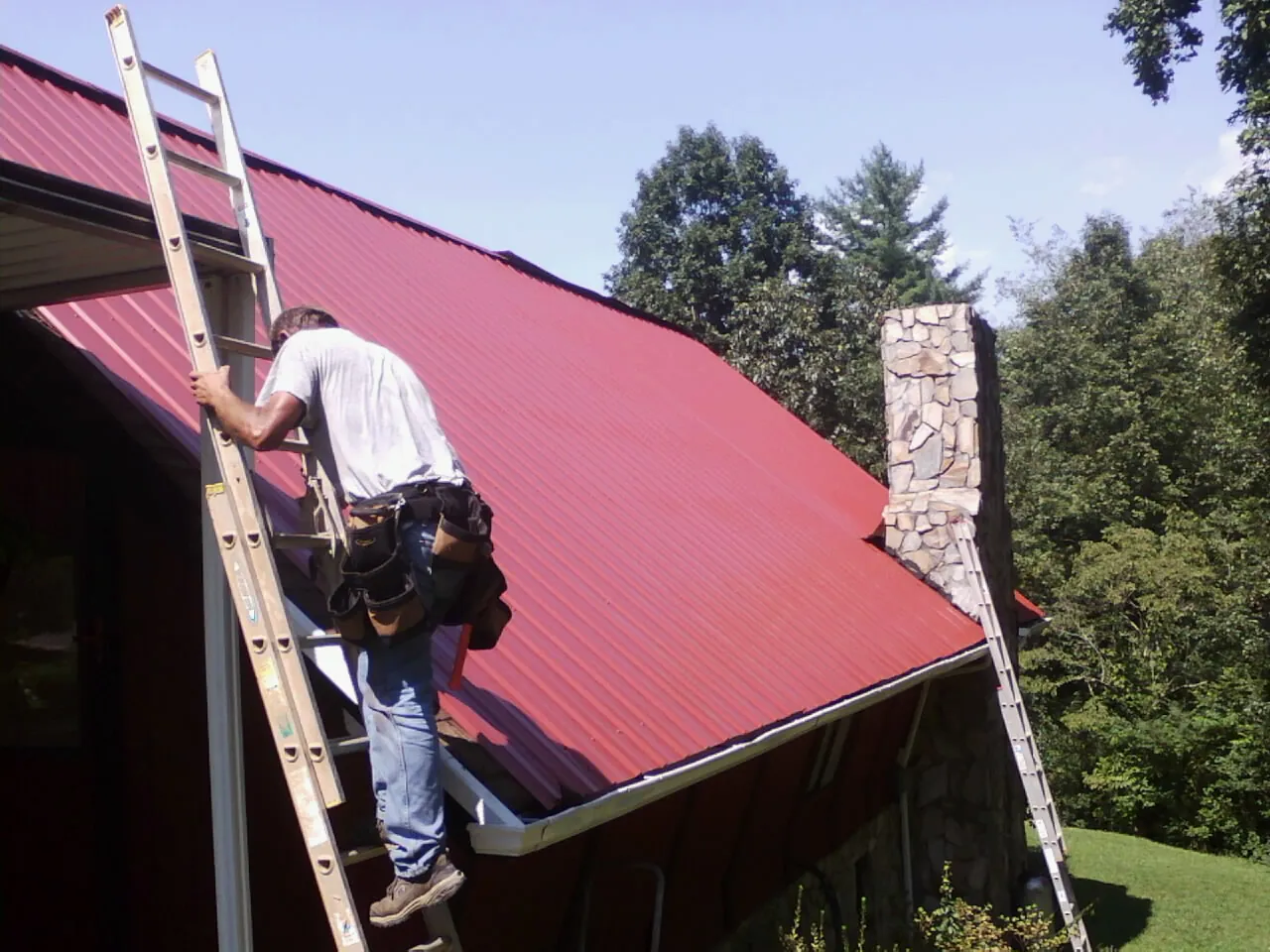 Roofer on a ladder beside a red metal roof and stone chimney — Soffit & Fascia Repair by Roofing Brothers in Jessie